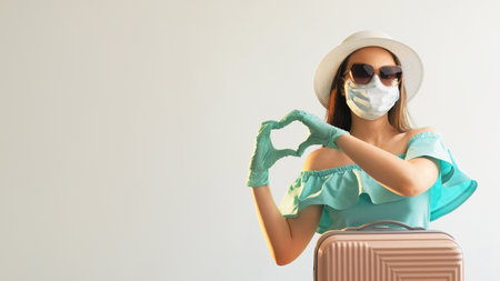 Pandemic love. Heart gesture. Girl in gloves mask summer dress sunglasses strew hat with suitcase showing hand affection sign isolated on white empty space background.の写真素材