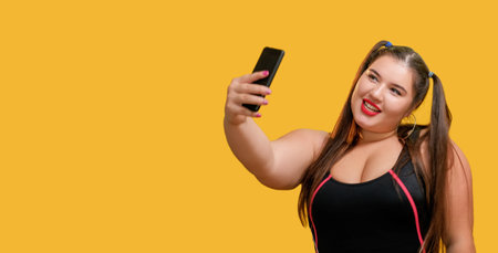 Making photo. Social media. Video blogging. Funny overweight chubby woman model taking selfie using phone streaming online isolated on yellow empty space background.の写真素材