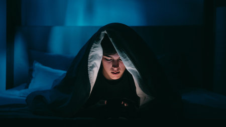 Illuminated by screen surrounded by calming blue ambient light man finds comfort hidden under a blanket using phone at night.の写真素材