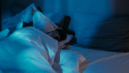 Pensive man trouble sleeping disturbed in bed with soft pillows under soothing dim blue light.の写真素材