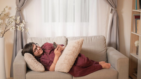 Phone entertainment. Home relax. Indoor leisure. Happy young woman typing on smartphone social media app laying resting on comfy couch in living room.の写真素材
