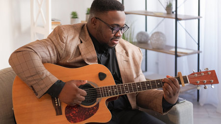 Male guitarist. Musical leisure. Melody practice. Talented satisfied man artist playing song chords on acoustic instrument on couch at home in living room.の写真素材