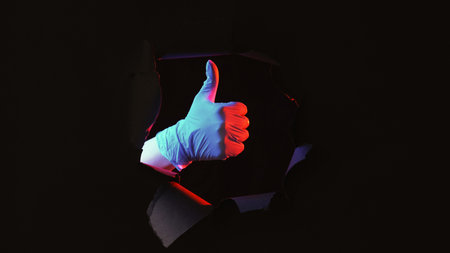 Doctor like. Experiment success. Red blue color light hand in glove approving with thumb up gesture in black breakthrough paper hole on dark empty space background.の写真素材