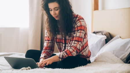 Home work. Freelancer woman. Online connection. Busy woman working online browsing laptop sitting on bed at light cozy bedroom.の写真素材