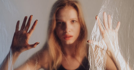 Female sorrow. Freedom desire. Disturbed woman trapped behind wrinkled transparent polyethylene film on gray out of focus background.の写真素材