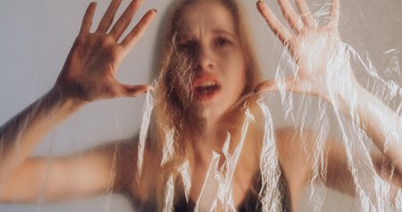 Breath lack. Asthma patient. Suffering woman trapped behind wrinkled transparent plastic polyethylene film isolated on gray out of focus background.の写真素材