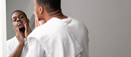 Skin routine. Beard care. Confident masculine man with white towel looking in mirror touching face stubble on light empty space background.の写真素材