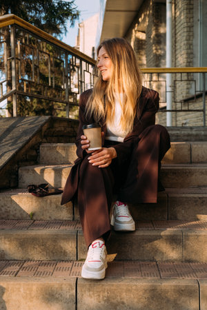 Urban weekend. Coffee leisure. Satisfied happy business woman enjoying sunlight sitting on stairs with hot drink in paper cup.の写真素材