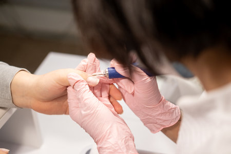 Hand manicure. Fingernails care. Female manicurist in protective pink gloves removing client nail plate with professional milling machine.の写真素材