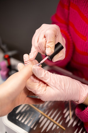 Nail procedure. Salon treatment. Beauty female master hands in protective pink latex gloves covering by acrylic varnish holding brush.の写真素材