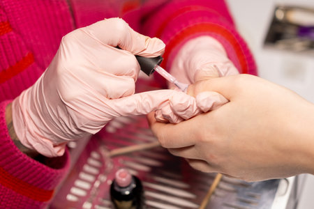 Hand manicure. Fingernails care. Female master in protective latex gloves applying decorative acrylic pastel varnish to nail plate with brush.の写真素材