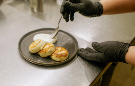 Cottage cheese pancakes. Sweet dish. Chef adding sour cream to portioned pancakes. Hands serving dessert plate on kitchen.の写真素材