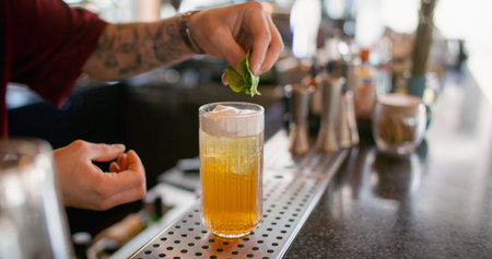 Serving drink. Barkeeper work. Tattooed hand adding green mint leaves to yellow refreshing cocktail...