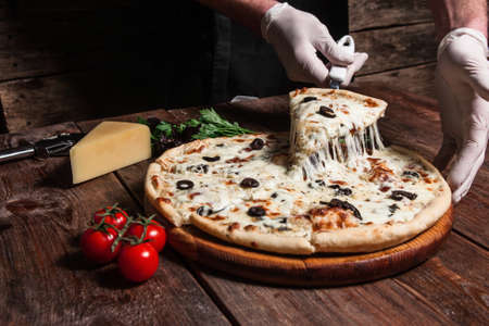 Chef lifting slice of hot tasty pizza with melting cheese on rustic wooden table. Italian cuisine, fast food.の写真素材