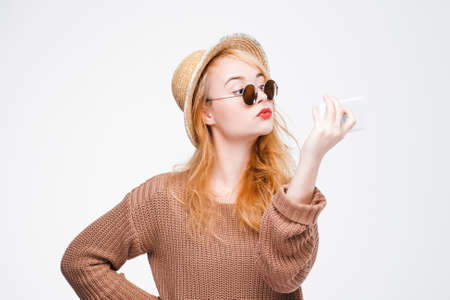 Nice young woman in straw hat and sunglasses taking selfie with mobile phone. Cool and fashionable hipster girl posing to smartphone camera. Grey background with copy space.の写真素材