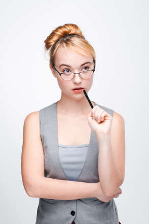 Young nice woman in glasses thinks of solving problem with pen near mouth, concentrated on finding right decision. Fair-haired thoughtful woman brainstorming on grey background.の写真素材