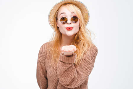 Young fashionable attractive hipster girl in sunglasses and straw hat sends air kiss to camera. Nice playful and happy woman with red lips on grey background with copy space.の写真素材