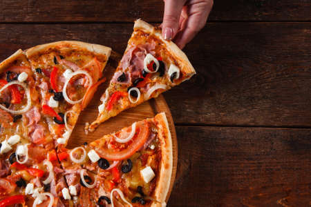 Hand taking hot slice of colorful delicious italian pizza served on wooden table, flat lay. Dark background with copy space.の写真素材
