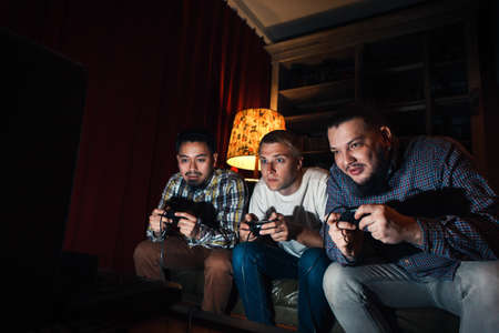 Three high concentrated young guys with joysticks play exciting video game at home in dark room. Tension to win tourney.の写真素材