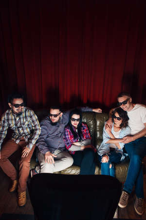 Group of young friends in 3d glasses have fun entertainment watching tv show on couch together, top view. Red curtain background with free space.の写真素材