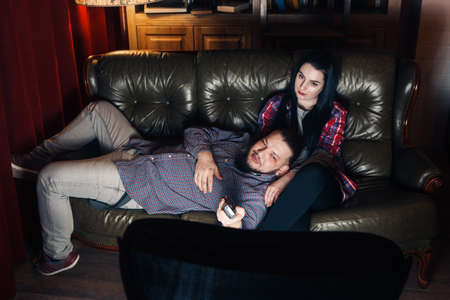 Couple of young tired people lay on couch and watch tv at home. Guy and girl bored from movie, changing channel with remote control.の写真素材