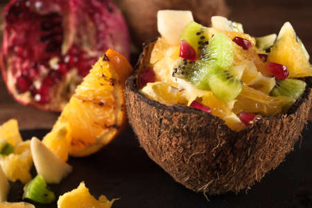 Juicy colorful fruit salad chopped in coconut peel on rustic wooden table with ingredients, close up...