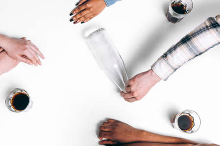 Four international friends play spin the bottle game. Interracial company, fun pastime, home leisure, first date concept. White table background with free space.の写真素材