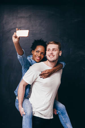 Man gives piggyback ride to woman, couple selfie on smartphone. Casual youth smiling and have fun on...