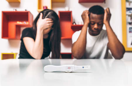 Positive test for pregnancy. Disappointment and frustrated young interracial couple, black man and white woman. Unplanned parenthood, think of abortion, safe sex.の写真素材