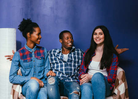 Happy company of three young friends together on pop blue background, international friendship. Smiling black man and two mixed races women sit at home on couch, fun pastime.の写真素材