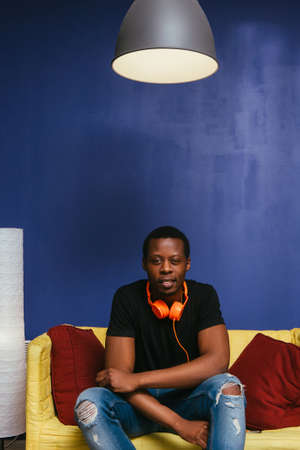 Young attractive black man sit on couch at home with orange headphones. Home leisure, pastime, lazy...
