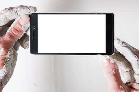 Unrecognizable worker with hands in white plaster hold mobile phone with empty screen, pov. Background of stucco wall with free space.の写真素材