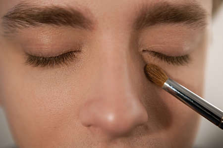 Applying concealer to cover dark circles under the eyes. Horizontal indoors close-up shot of man with eyes closed and brush for makeup. Male beauty , gender equality concept.の写真素材