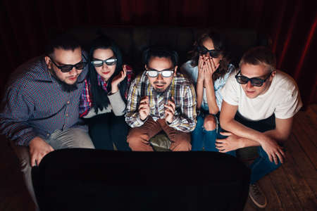 Young people in 3d glasses watch tv on couch. Five friends amazed by exciting effects of new...