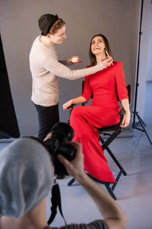 Visagist do makeup for happy woman on photoshoot. Photographer and stylist work in studio . Creative team make commercials. Photo school, lookbook, fashion backstage conceptの写真素材
