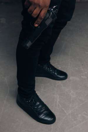 Unrecognizable black criminal with gun closeup studio shoot. Armed gangster man on dark background. Outlaw, ghetto, murderer, robbery conceptの写真素材