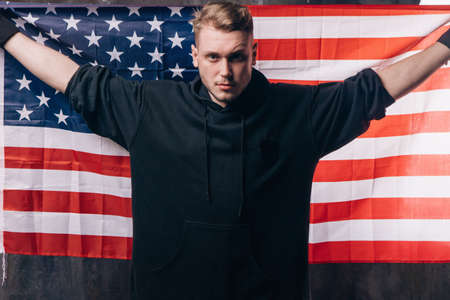 Young USA patriot holds national flag. Strong man in black cloth with stars and stripes behind him. Independence day, confidence, pride, fidelity to the nation, memorial day conceptの写真素材