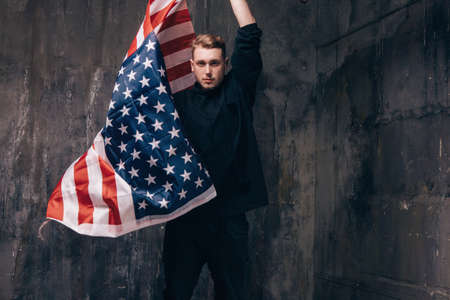 Young USA patriot with flying national flag. Strong man in black cloth on dark studio background. Independence day, confidence, pride, fidelity to the nation, memorial day conceptの写真素材
