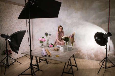 Decorator make flower bouquet at studio. Young girl makes an ornament from white flowers in a workshop. Photoshoot backstageの写真素材