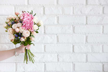 Female hand show bouquet with rose flowers on white brick background. Gift for mother or woman, florist work, wedding decor, beautiful bouquet sale conceptの写真素材
