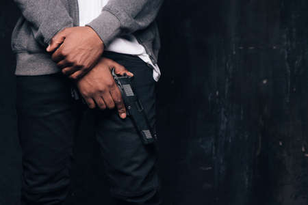 Unrecognizable armed black criminal man studio shoot. Gangster guy with gun in hand on dark background with free space. Outlaw, ghetto, murderer, robbery conceptの写真素材