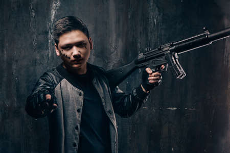 Tattooed face guy with sniper rifle pointing. Armed asian killer man with weapon and tattoo on dark...