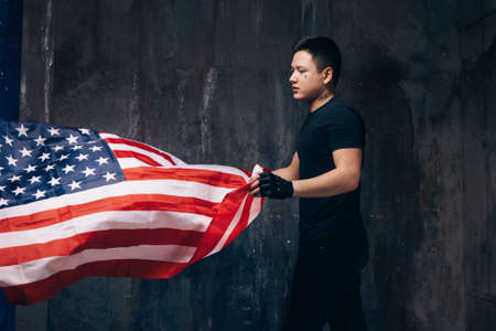Young USA patriot with flying national flag. Strong man in black cloth with tattoo on dark studio background. Independence day, confidence, pride, fidelity to the nation, memorial day conceptの写真素材