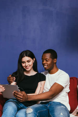 Happy smiling international couple sit with tablet on couch at home. Surfing internet, watch photos and movie. Togetherness, happiness, relationship, leisure concept.の写真素材