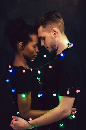 Interracial young couple tied together with sparkling garland colorful lights. Togetherness, tender, love, new year holiday, happy relationship and unity concept.の写真素材