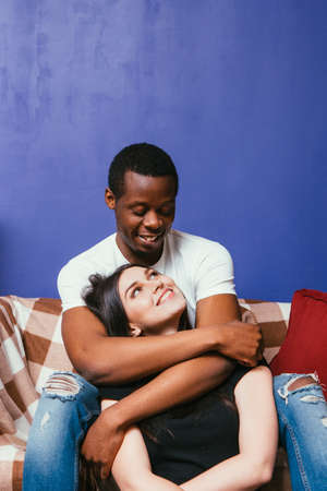 Young happy couple in love. Black man and white woman cuddle and caress on couch at home. Happiness, joy, hugs, international relationship concept.の写真素材
