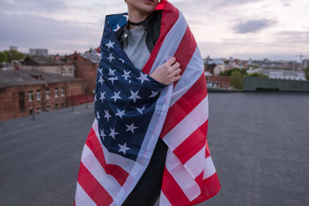 Girl dressed in USA flag on city and sky background. Unrecognizable american patriot, national event celebration, pride, usa citizen conceptの写真素材
