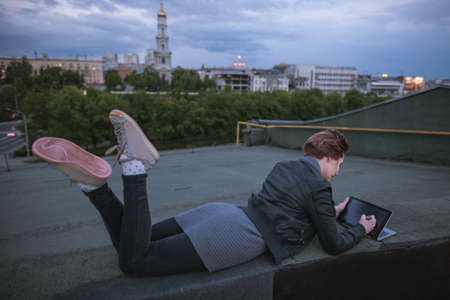 Designer with tablet is drawing on cloudy sky background. Creative woman lying on the buildings roof in city during work. Blogger, distant work, wireless internet , art profession conceptの写真素材