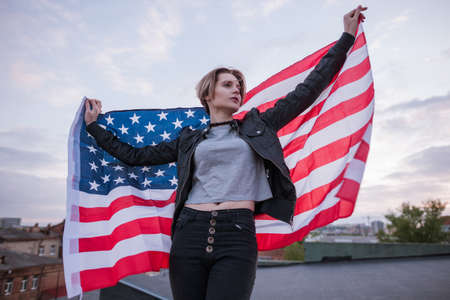 Young girl with USA flag behind the back on evening sky background. Patriot, national event celebration, pride, usa citizen conceptの写真素材