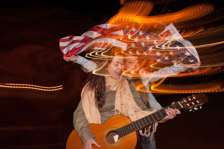 Girls celebrate Independence Day at night. Singer with guitar and happy girl with flying american flag on blurred city lights background. Youth lifestyle conceptの写真素材
